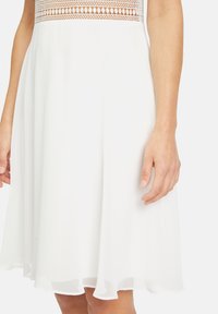 Vit klänning med en åtsittande spetsliv och flödande kjol, med en fåll som mjukt svänger ut. Tyget verkar vara lätt och texturerat.