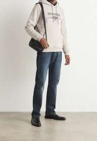 Sudadera beige clara con logo impreso, jeans de denim azul y zapatos de cuero negros. Accesorio: bolso cruzado negro con detalles texturizados.