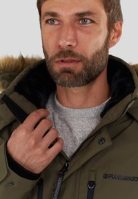 Uomo con barba che indossa una giacca invernale verde oliva con cappuccio foderato in pelliccia, mentre sistema il colletto sopra un maglione grigio.