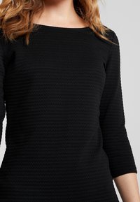 Musta mekko, jossa on teksturoitu, sahalaitakuvio, sekä veneen kaula-aukko ja kolmannen neljänneksen hihat. Kangas näyttää pehmeältä ja hieman venyvältä.