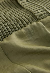 Gros plan d’un tissu vert olive combinant des textures en maille côtelée et un tissage lisse dans un design superposé.