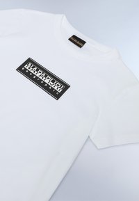 Valge puuvillane T-särk lühikeste varrukatega, millel on must ristkülikukujuline logo, mis kannab valget tekstikirja "NAPAPURI geographic".