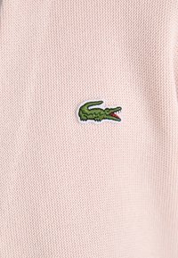 Um tecido de malha rosa apresenta um logótipo de crocodilo bordado em verde. A textura parece bem ajustada, com um padrão subtil.