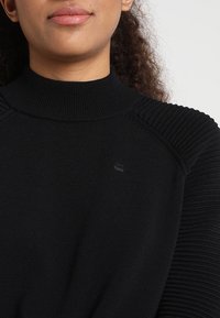 Svart stickad tröja med hög krage, med ribbade axeldetaljer och en liten logotyp på bröstet. Slät textur genom hela plagget.