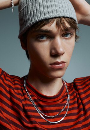 Fiatal személy kék szemekkel, szürke kötött sapkát, piros-fekete csíkos inget visel, és két ezüst nyaklánca van, kezével a sapkát igazítja.