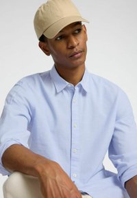 Camicia azzurra chiara con maniche lunghe, realizzata in un tessuto morbido. Cappello beige con visiera arrotondata, posizionato in modo inclinato.