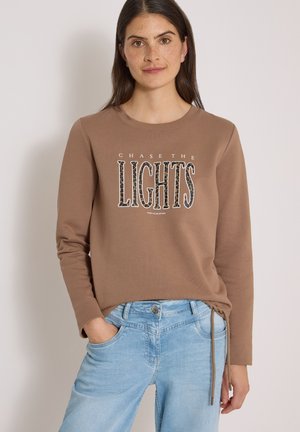 Kobieta z długimi ciemnymi włosami ubrana w brązową bluzę z napisem "CHASE THE LIGHTS" i jasnoniebieskie dżinsy, stojąca z jedną ręką w kieszeni.