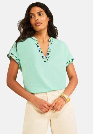 Frau mit langen dunklen Haaren, die ein mintgrünes Oberteil mit floralem Besatz am V-Ausschnitt und an den Ärmeln, cremefarbene Hosen und goldene Armbänder trägt.