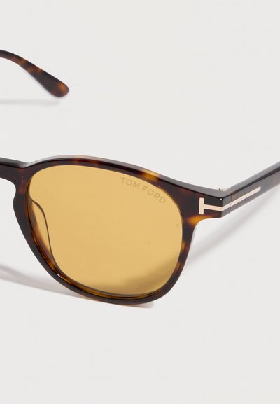 Καφέ γυαλιά ηλίου με σχέδιο χελωνόστρακα, με κεχριμπαρένιους φακούς και χρυσά διακοσμητικά λογότυπα "T" στις πλευρές, τοποθετημένα σε λευκή επιφάνεια υπό γωνία.