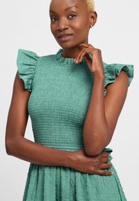 Mintgrünes Kleid aus strukturiertem, gerafftem Stoff, mit Rüschenärmeln und hohem Kragen sowie einem ausgestellten Rock.