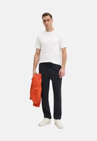 T-shirt bianco con logo in rilievo, pantaloni della tuta blu scuro con cordino, e felpa arancione. Modello che indossa sneakers bianche. Outfit semplice e casual.