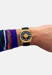 Achtkantige goldene Uhr mit schwarzem Gummiband, dunkelblauem Zifferblatt mit weißen Ziffern und einem Logo, montiert an einem Handgelenk mit einem bunten Pullover.