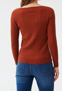 Femme portant un pull en maille torsadée couleur rouille et un jean bleu, vue de dos sur un fond blanc uni.