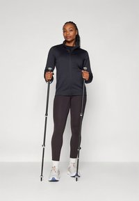 Fekete cipzáras dzseki, testhez simuló sötét leggings, túrabotok; a modell fehér sportcipőt visel színes részletekkel. Egyszerű háttér.