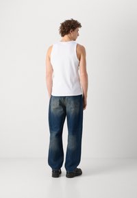 Débardeur blanc sans manches, jeans bleu foncé décontractés avec un effet délavé, et baskets noires. Vu de dos, montrant les détails du vêtement.