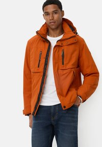 Pomarańczowa wodoodporna kurtka z kapturem, z zamykanymi kieszeniami i fakturowanym wykończeniem. Zestawiona z białą koszulą i ciemnoniebieskimi dżinsami.