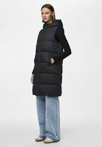 Gilet imbottito nero con cappuccio, design trapuntato e tasche laterali. Abbinato a jeans larghi azzurri e sneaker beige.
