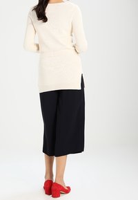 Cremfarbener Longsleeve-Strickpullover mit Seitenschlitzen, kombiniert mit navyblauen Culottes und roten Wildleder-Blockabsatzschuhen, von hinten betrachtet.