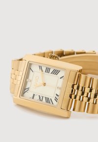 Χρυσό ορθογώνιο ρολόι με λευκό καντράν, μαύρους ρωμαϊκούς αριθμούς και δύο μικρούς κρυστάλλους. Διαθέτει ατσάλινη βραχιόλι με υφή σε χρυσό χρώμα.