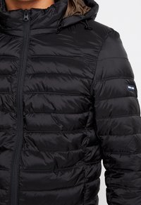 Schwarze Pufferjacke mit gestepptem Design, Kapuze und seitlichen Taschen. Verfügt über einen Frontreißverschluss und ein Logo-Patch am Ärmel. Glatte Textur.