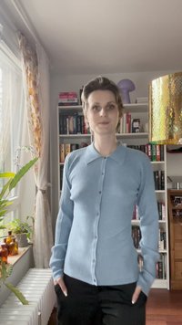 Ανοιχτό μπλε ριγέ κασκόλ με γιακά, διαθέτει κουμπιά μπροστά και μακριά μανίκια. Το μοντέλο στέκεται σε ένα δωμάτιο με ράφια γεμάτα βιβλία.