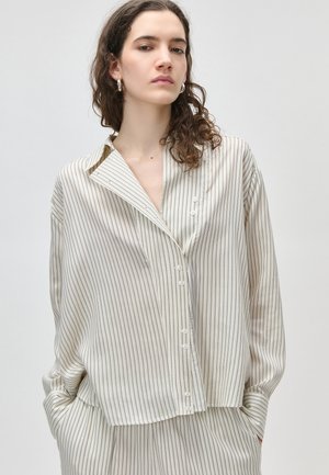 Joven con cabello rizado, vistiendo una blusa crema de gran tamaño y pantalones a rayas a juego, de pie con las manos en los bolsillos sobre un fondo liso.