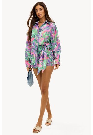 Langarmshirt met levendig tropisch bladerenpatroon, met tinten groen, roze en paars. Geknoopt in de taille, gecombineerd met spijkerbroek en sandalen.