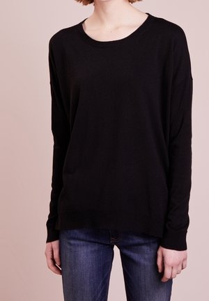 Femme portant un pull noir uni à manches longues et un jean bleu, debout devant un fond rose clair uni.