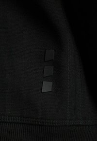 Tela negra con un tejido texturizado. Presenta tres pequeños cuadrados negros como acentos a lo largo de la costura. Diseño limpio y minimalista sin patrones.