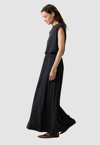 Zwarte, mouwloze maxi-jurk met een elastische taille, vloeiende stof en een gladde textuur. Geaccentueerd met een gouden armband en patroonballetjes.