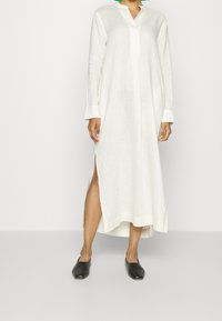 Robe longue blanche en lin avec col et patte de boutonnage devant, fentes latérales et manches longues. Associée à des chaussures noires faciles à enfiler.
