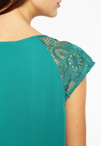 Groenblauw top van gladde stof met fijn kanten bies op de schouders, met bloemige en geometrische patronen in een contrasterende kleur.