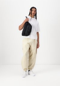 Mala sling preta com fecho, usada por um modelo com uma t-shirt branca e calças bege, de pé sobre um fundo branco.