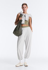 Mlada ženska, oblečena v olivno zelen kratki top, svetlo sive trenirke, bele superge, sivo jopico čez ramena, drži vrečko z leopardjim vzorcem.