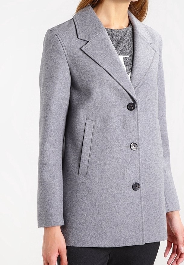 Femme portant un manteau en laine gris clair à simple boutonnage avec trois boutons noirs et des poches latérales, par-dessus un haut gris.