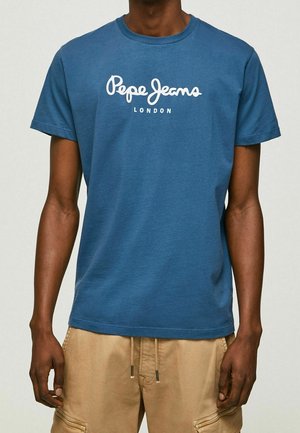 Homme portant un t-shirt bleu Pepe Jeans London et un pantalon beige, debout sur un fond clair uni.