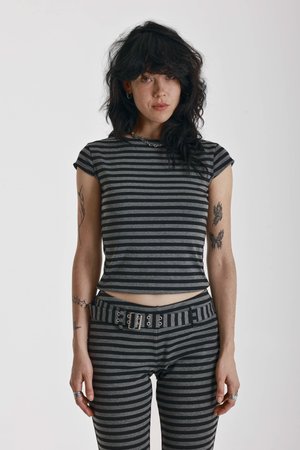 Femme aux cheveux noirs ondulés portant une chemise rayée noire et grise et un pantalon assorti avec une ceinture, debout devant un fond blanc uni.
