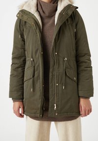Olijfgroene parka jas met een nepbont gevoerde capuchon, ritssluiting en drukknopen, verstelbare trekkoorden en twee voorkeurs.