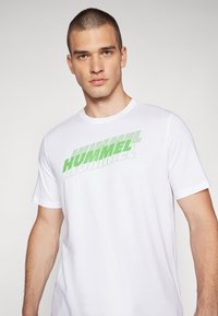 Valge puuvillane T-särk, millel on suure rohelise graafilise teksti "HUMMEL" rindkere kohal, lühikesed varrukad ja ümmargune kaelus. Sujuv tekstuur.
