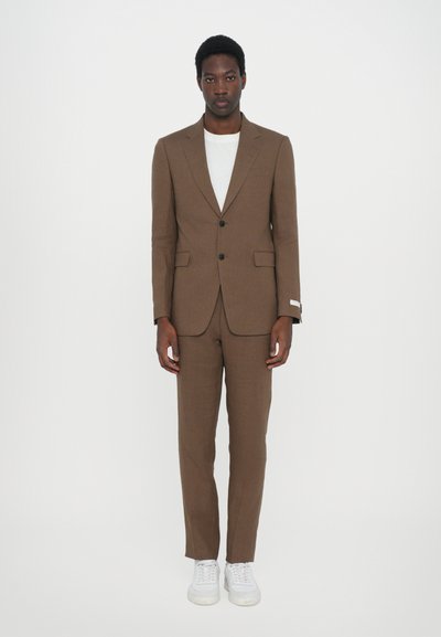 Costume marron sur mesure avec une coupe ajustée, veste à un bouton, poches plaquées et pantalon assorti. T-shirt blanc et baskets blanches portés.