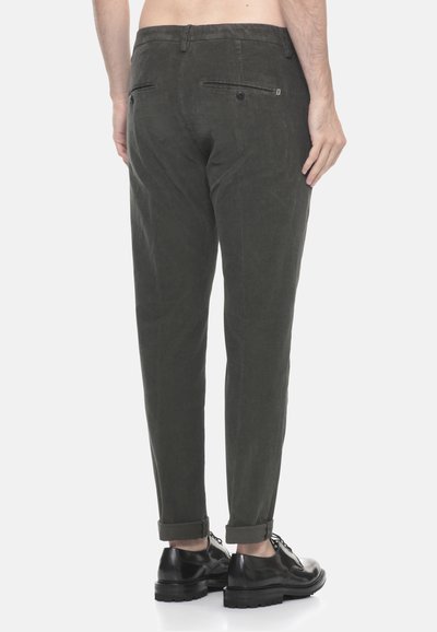 Uomo che indossa pantaloni di velluto a coste verde scuro con orli risvoltati e scarpe eleganti in pelle nera, in piedi contro uno sfondo neutro.