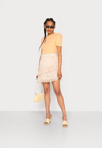 Top laranja de mangas bufantes com uma saia creme em camadas e florais. Combinado com mules amarelas e uma pequena mala amarela. Óculos de sol pretos completam o visual.