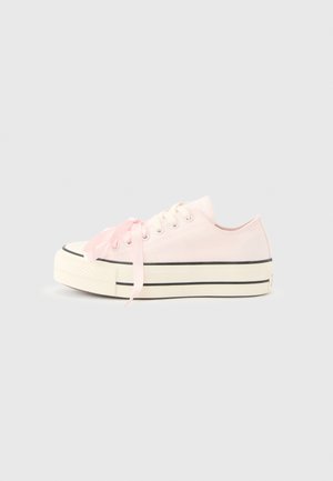 Baskets basses rose pastel avec semelle blanche, bandes noires et larges lacets en satin sur un fond gris clair uni.