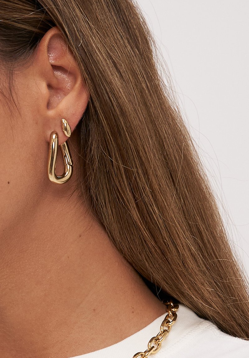 Nahaufnahme eines Ohres mit großen goldenen Creolen und kleinem goldenen Stecker, hellbraunem Haar und einer massiven goldenen Kette auf einem weißen Oberteil.