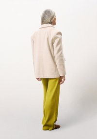 Blazer en laine beige avec un col cranté, associé à un pantalon large jaune vif et des chaussures marron, vu de dos.