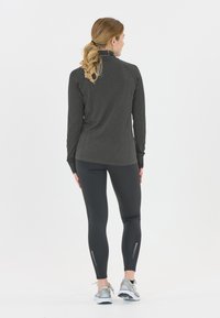 Frau mit blonden Haaren im Pferdeschwanz trägt ein dunkles Langarmoberteil, schwarze Leggings mit reflektierenden Streifen und graue Laufschuhe, steht mit dem Rücken zur Kamera.