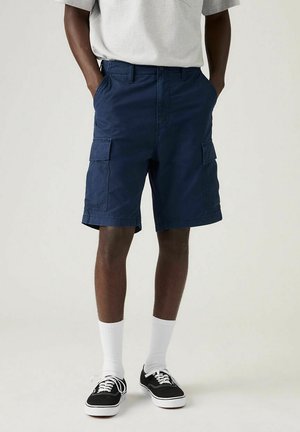 Homme portant un short cargo bleu marine, des chaussettes blanches montantes, des baskets noires et blanches et un t-shirt gris clair, debout les mains dans les poches.