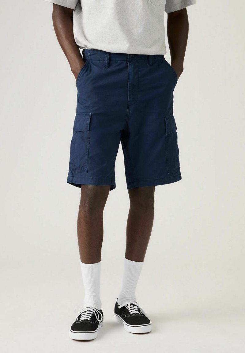 Homme portant un short cargo bleu marine, des chaussettes blanches montantes, des baskets noires et blanches et un t-shirt gris clair, debout les mains dans les poches.