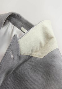 Grå kordflossad tyg med en filtkrage. Kragen har en krämfärgad nyans och synliga stygn. Det inre fodret är vitt, vilket visar struktur och textur.