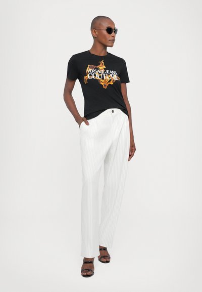T-shirt graphique noir avec impression jaune et orange et pantalon à rayures blanches. Le modèle porte des lunettes de soleil noires et des sandales noires à lanières.